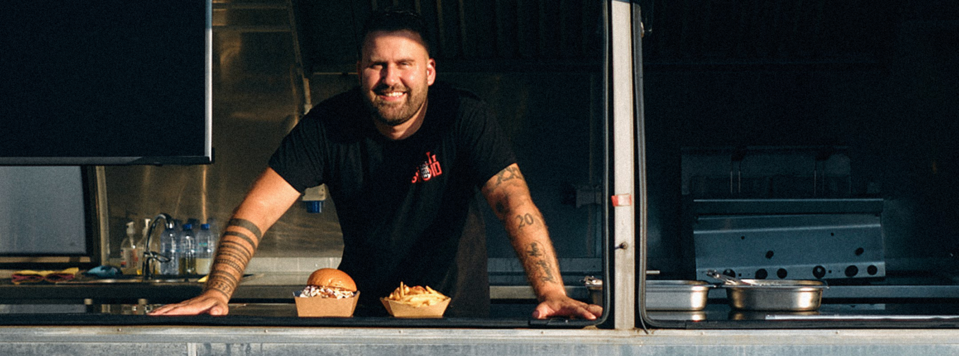 Χαμογελαστός σεφ ακουμπά στον πάγκο του food truck με μπέργκερ και πατάτες