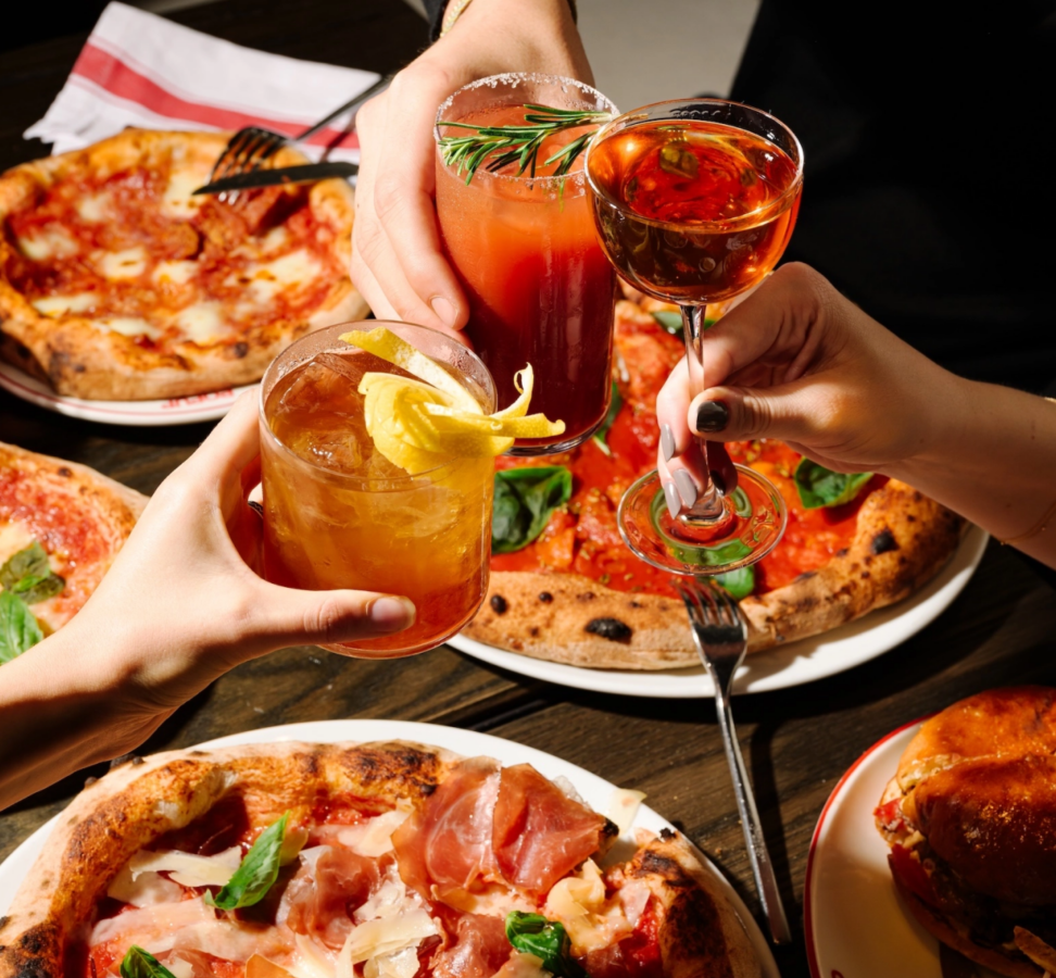 Hands toasting cocktails over a table of pizza